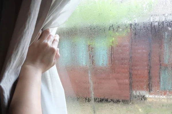 Woman's hand opening curtains in the bedroom with rain droplet on the glass, natural light and blurred garden background with text space