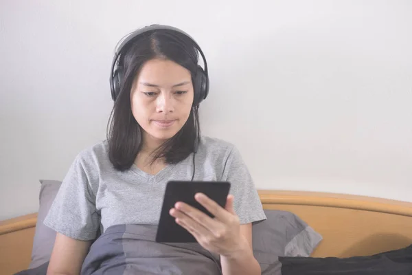 アジアの女性の美しいヘッドフォンとベッドでリラックスの 読者と電子本を読んで音楽を聴いて ストック画像