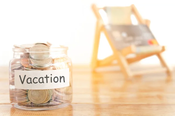 Vacation budget concept. Holidays money savings concept. Collecting money in the money jar for Vacation. Money jar with coins and beach seat on wooden background.