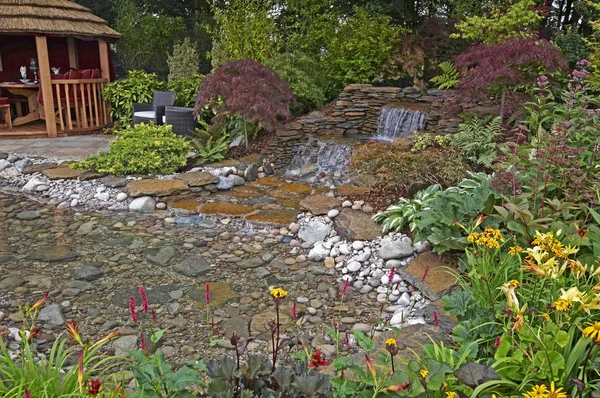 Водний сад з водоспадом в лісовій місцевості з квітами, і розслабляючий простір з барвистими квітами і сидіннями — стокове фото