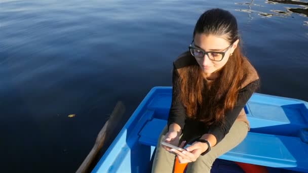 Hermosa chica sentada en un barco con un teléfono. En cámara lenta. Hermosas olas en el agua — Vídeo de stock