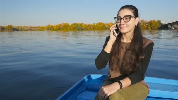 La fille parle au téléphone alors qu'elle est assise dans un bateau sur l'eau. Joli sourire. Journée ensoleillée. Mouvement lent — Video