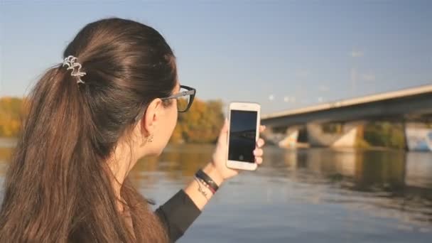 Menina bonita tira fotos da natureza. Rio com uma ponte. A paisagem pitoresca. A filmar ao telefone. Movimento lento — Vídeo de Stock