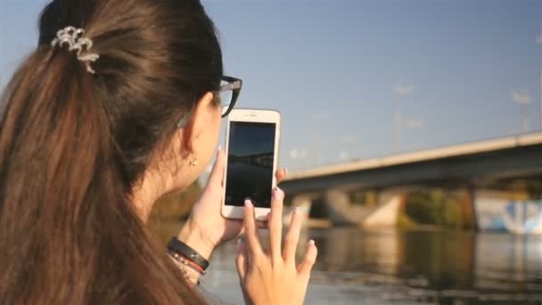 若い女の子が電話で写真を撮ります。川に美しい橋があります。絵のように美しい風景。クローズ アップ。晴れた日 — ストック動画