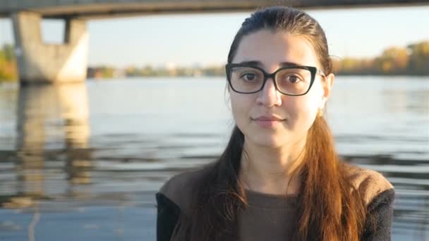 Menina bonita em óculos está olhando para a câmera e sorrindo. Sorriso adorável. Contra o fundo do rio com uma ponte. Um bom lugar. dia ensolarado no verão — Vídeo de Stock