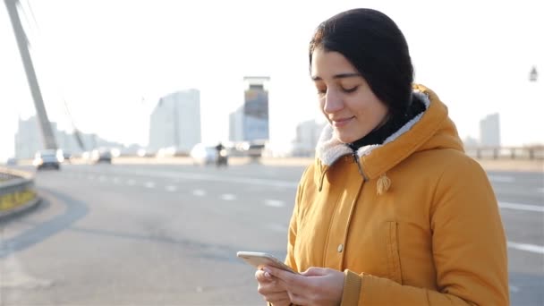 Menina bonita escreve uma mensagem em um smartphone. Movimento lento. Estrada em segundo plano — Vídeo de Stock