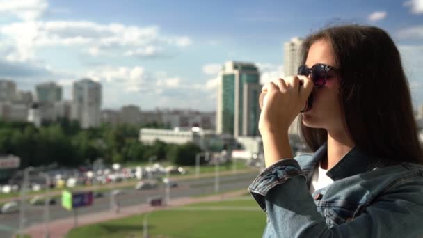 Glimlachend brunette meisje drinkt koffie tegen stadsgezicht traag — Stockvideo