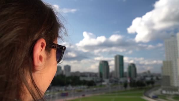 Брюнетка в сонцезахисних окулярах захоплюється міським пейзажем під хмарним небом — стокове відео
