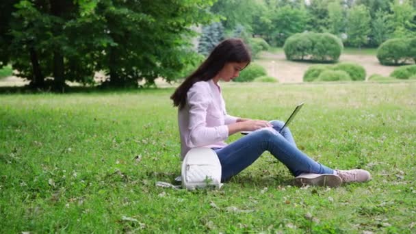 Langharige brunette zit op groen gras en typen op laptop — Stockvideo
