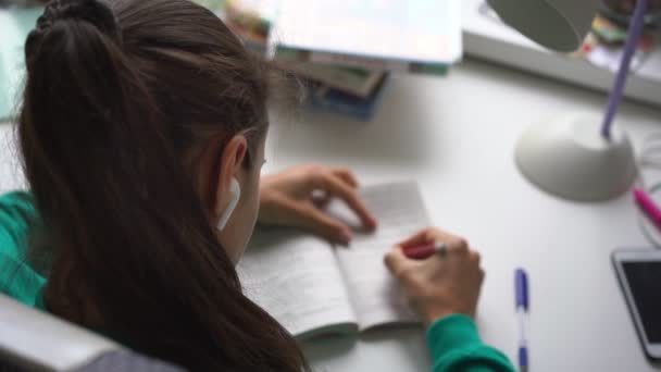 Meisje tiener student in draadloze oortelefoons schrijft thuis taak — Stockvideo