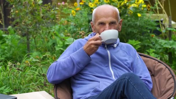 Ανώτερος άνθρωπος πίνει τσάι σε ζεστή πολυθρόνα στο ανθισμένο κήπο — Αρχείο Βίντεο