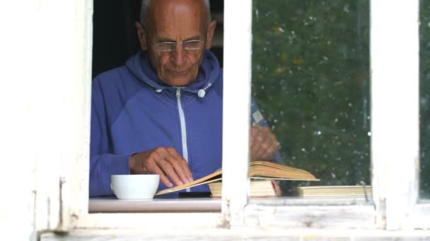Старик читает книги и пьет чай на подоконнике крупным планом — стоковое видео