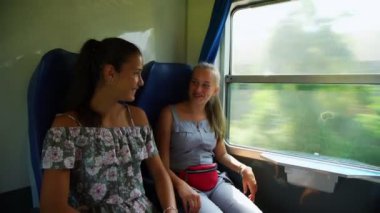 Kız arkadaşlar trende gülümseyip pencereden dışarı bakarlar.