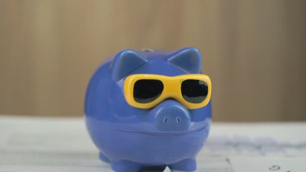 Münzen fallen auf blaues Sparschwein mit Sonnenbrille — Stockvideo