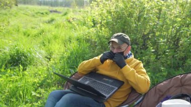 Maskeli emekli bahçede dizlerinin üstünde dizüstü bilgisayar tutuyor.