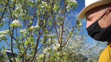 Şapkalı ve siyah maskeli emekli çiçek açmış ağacın yanında duruyor.