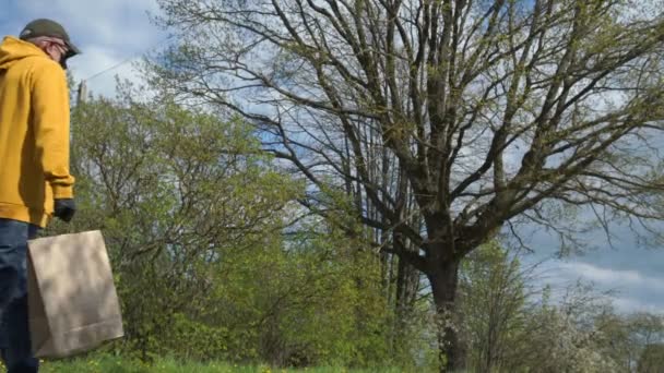 Senior man in gele hoodie draagt papieren zak langs bomen — Stockvideo