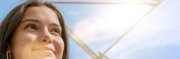 Smiling young woman with long hair against amusement park attraction at back bright summer sunlight close low angle shot