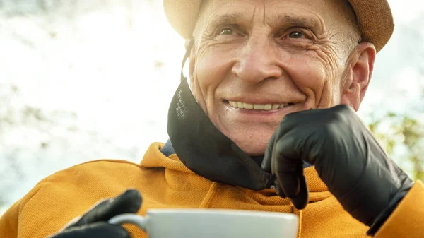 Старий в чорній стерильній масці і рукавичках посміхається тримаючи чашку — стокове фото