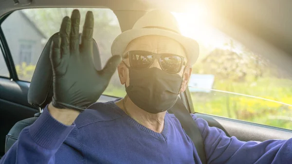 Maskeli kıdemli sürücü arabada oturuyor ve eldiven takıp el sallıyor.
