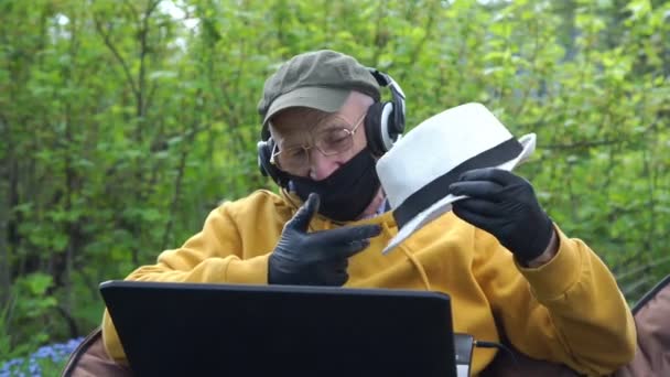 Pensionista en máscara desechable y auriculares muestra sombrero blanco — Vídeos de Stock