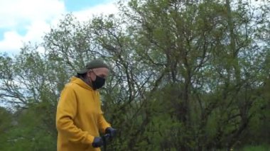 Maskeli son sınıf öğrencisi mobiletiyle yeşil ağaçların yanından geçiyor.