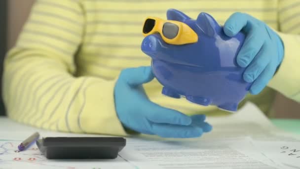 Armer Mann in blauen sterilen Handschuhen schüttelt Sparschwein langsam — Stockvideo