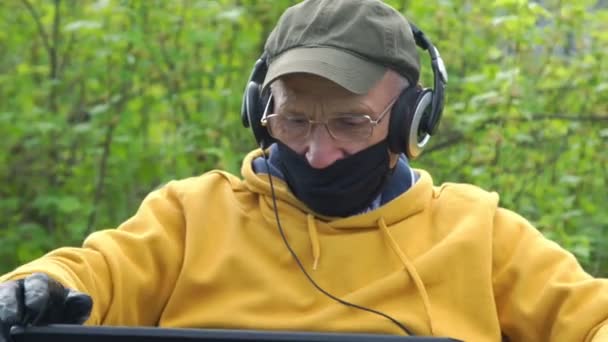 Старик в маске и наушниках типов на ноутбуке в саду — стоковое видео