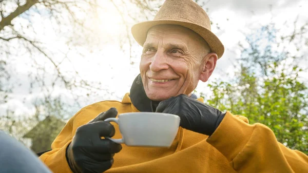 Старший чоловік в жовтому светрі і капелюсі посміхається тримаючи чашку — стокове фото