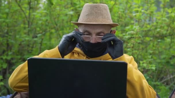 Pensioner em chapéu e máscara coloca em óculos segurando laptop — Vídeo de Stock