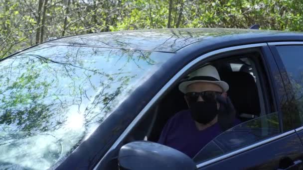 Alter Mann mit schwarzen Maskenhandschuhen sitzt im Auto und winkt mit der Hand — Stockvideo