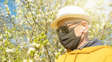 Yüzü maskeli ve kulaklıklı son sınıf öğrencisi çiçek açmış ağacın yanında.