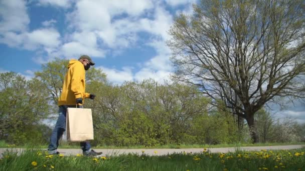 Emeryt w żółtej bluzie niesie torbę z jedzeniem do domu przez drzewa — Wideo stockowe
