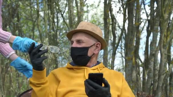 Старик в маске и перчатках берет очки против сада — стоковое видео