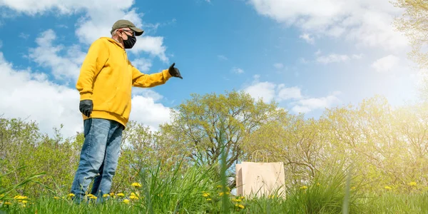 Alter Mann mit Maskenhandschuhen zeigt auf Tasche im Gras unter blauem Himmel — Stockfoto