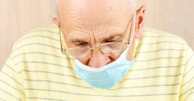 Mavi tek kullanımlık maskeli ve gözlüklü yaşlı adam aşağı bakıyor.