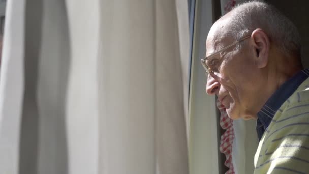 Лысый старик в очках смотрит в окно через занавески — стоковое видео