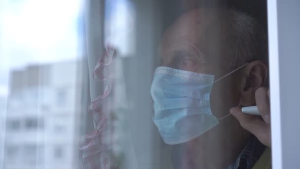 Hombre viejo triste en la máscara mira fuera de la ventana vista a través de vidrio — Vídeos de Stock