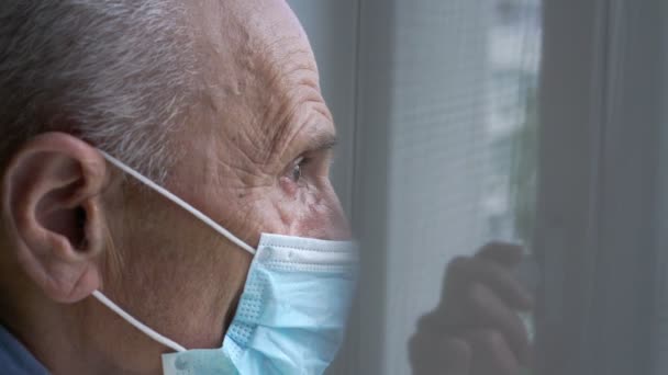 Senior man with wrinkled face in blue facial mask at window — Stock Video