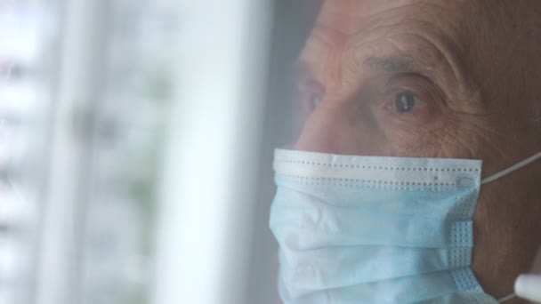 Sad pensioner in blue facial mask looks straight by window — Stock Video