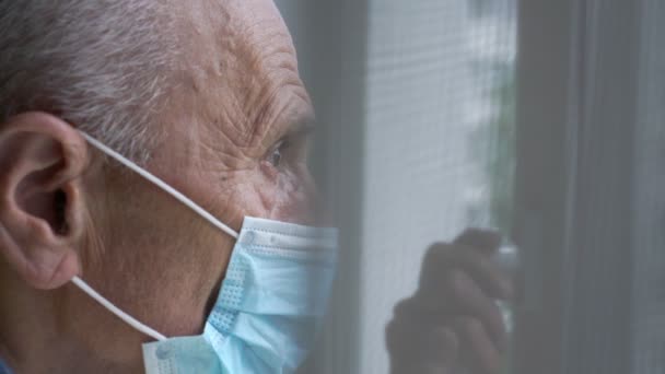 Oude man in wegwerp masker kijkt naar stadsgezicht door raam — Stockvideo