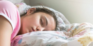 Tişörtlü kız büyük yatakta uyur, yastığa sarılır.