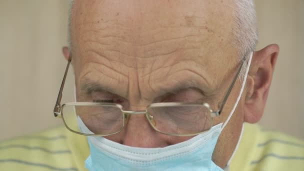 Stárnoucí člověk v brýlích a modré sterilní masce se dívá dolů — Stock video
