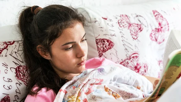 young woman with ponytail lies on large bed and reads book