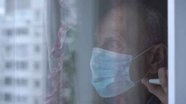 Wieku mężczyzna w niebieskim jednorazowej maski stoi za oknem — Wideo stockowe
