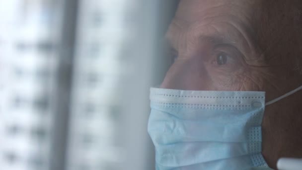 Pensionista deprimido en azul mascarilla desechable en la ventana — Vídeos de Stock