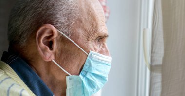 Maskeli gri saçlı adam pencerenin kenarında dikiliyor.