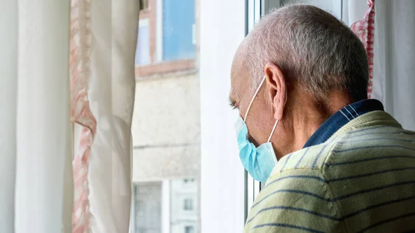 Mavi steril maskeli yaşlı adam pencerenin yanında duruyor.