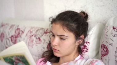 Genç esmer kadın yatakta büyük bir yastığa uzanıp kitap okuyor.