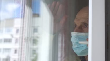 Mavi tek kullanımlık maskeli kıdemli adam pencerede el ele dalgalanıyor.